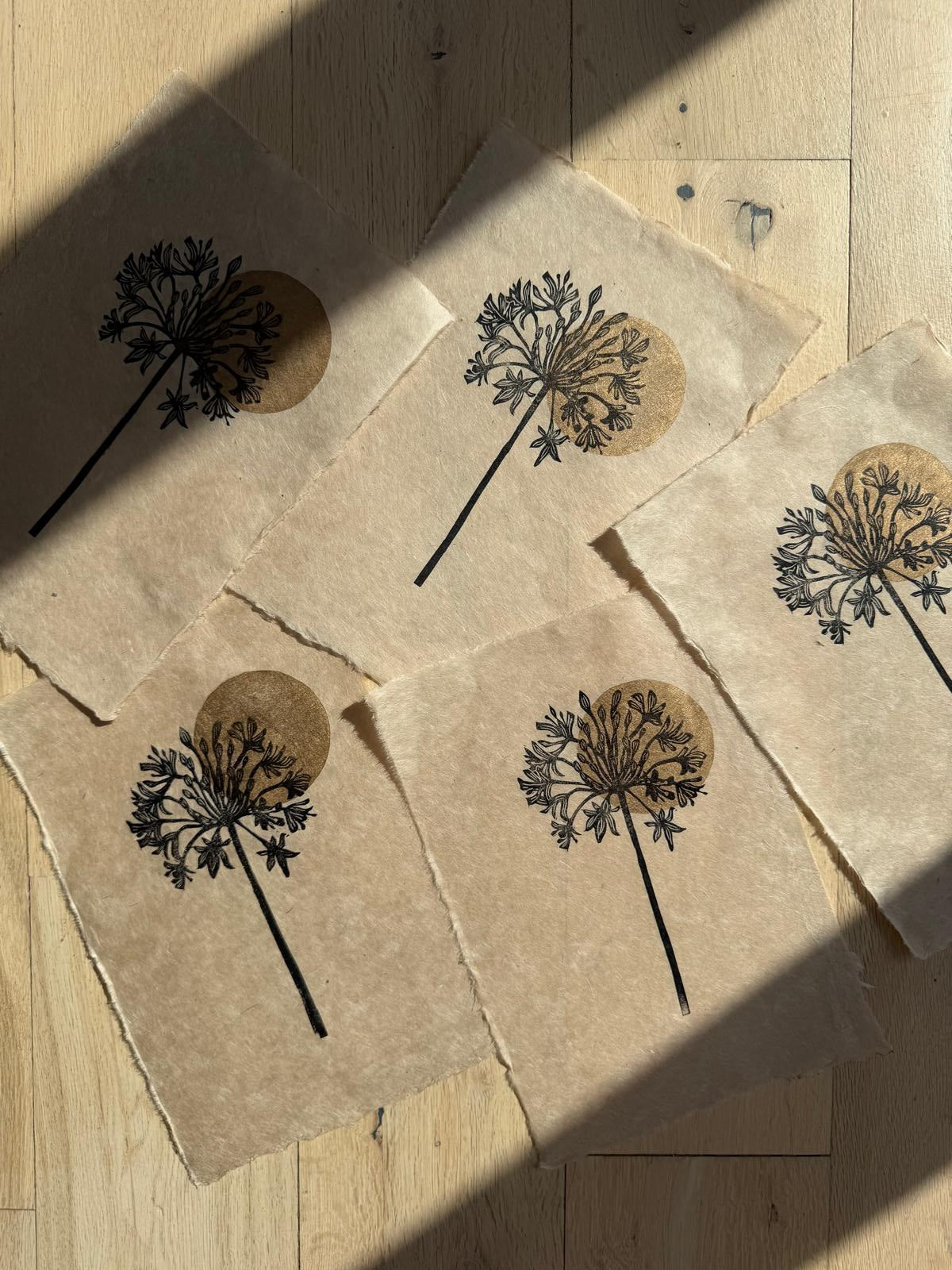 Five square pieces of textured beige paper with torn edges, each printed with a black agapanthus design and a circular gold patch, arranged on a light wood floor under diagonal shadows.
